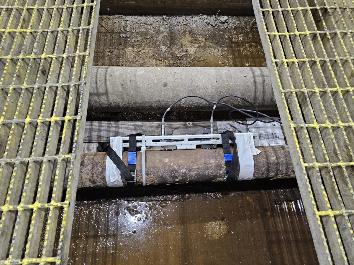 Clamp-on ultrasonic transducer rail strapped to a process pipe beneath walkway grating in a treatment plant